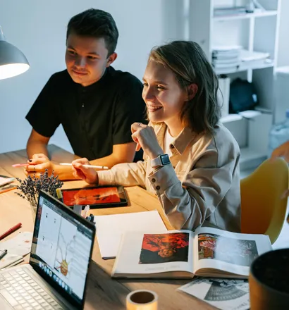 Deux jeunes étudiants collaborant sur un projet à une table avec un ordinateur portable, une tablette et un livre ouvert.